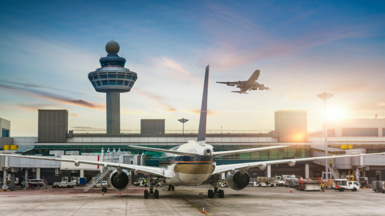 A large airplane is parked on an airport tarmac with a control tower in the background. Another plane is in the sky, preparing to land. The scene is set against a vibrant sunset.