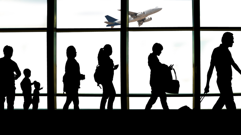 Silhouette of people walking in an airport terminal against a large window, with an airplane taking off in the background.