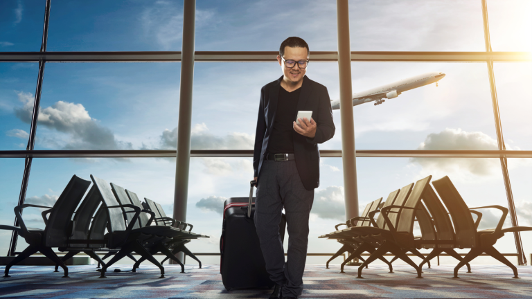 A man with glasses, wearing a black blazer, stands in an airport terminal holding a smartphone. He is pulling a suitcase. The sun shines through large windows, and a plane is visible in the sky outside. Empty seats line the terminal.