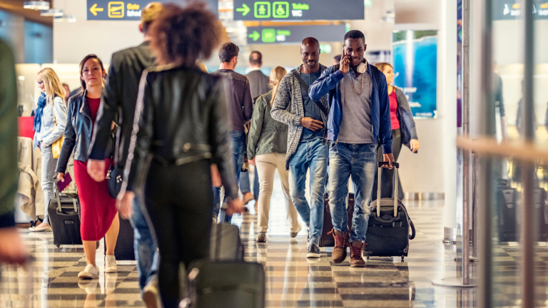 A busy airport scene with travelers walking through a terminal. People are carrying luggage, checking their phones, and wearing casual attire. Overhead signs indicate directions. The environment is bright and spacious.