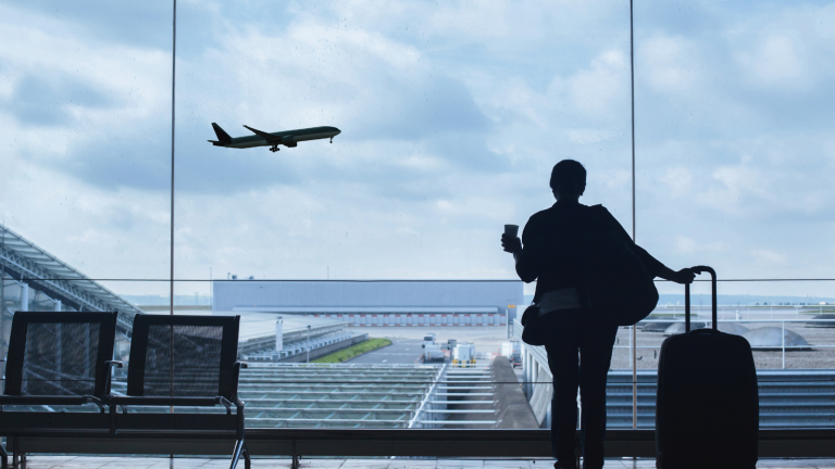 A person stands by a large airport window, holding a cup and luggage, watching a Sun City transfer plane take off. The scene is set against a cloudy sky, with an airport runway and terminal visible in the background.