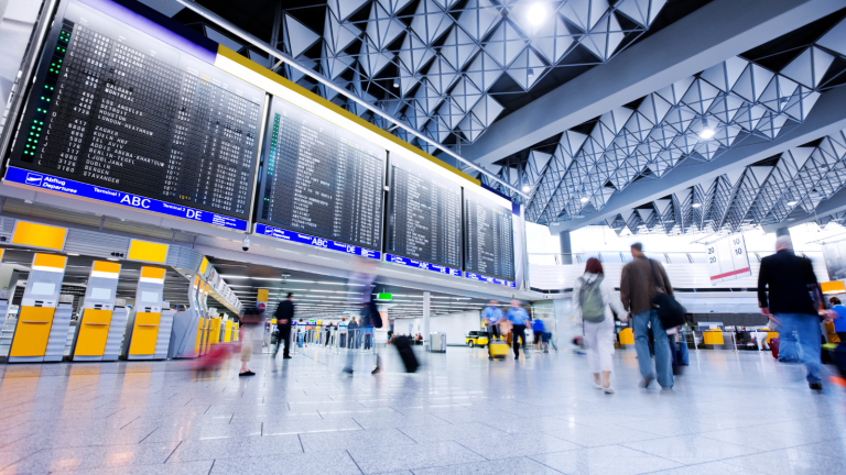 A bustling airport terminal hums with travelers walking and rolling luggage, assisted by Sun City Transfers. Large digital flight boards display arrivals and departures beneath geometric ceiling patterns and bright lighting.