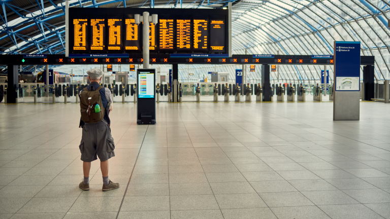 A person wearing a backpack stands in a spacious train station, gazing at the large electronic departure board. The Sun City Transfers logo glints under a high, glass-paneled ceiling, with rows of ticket barriers visible in the background.