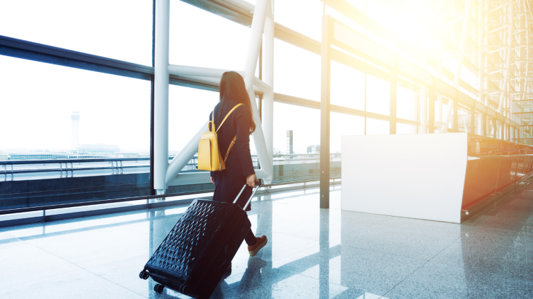 A person with long hair walks through an airport terminal, pulling a large black suitcase. They wear a yellow backpack and dark clothing. Sunlight streams through the large glass windows, illuminating the spacious interior.