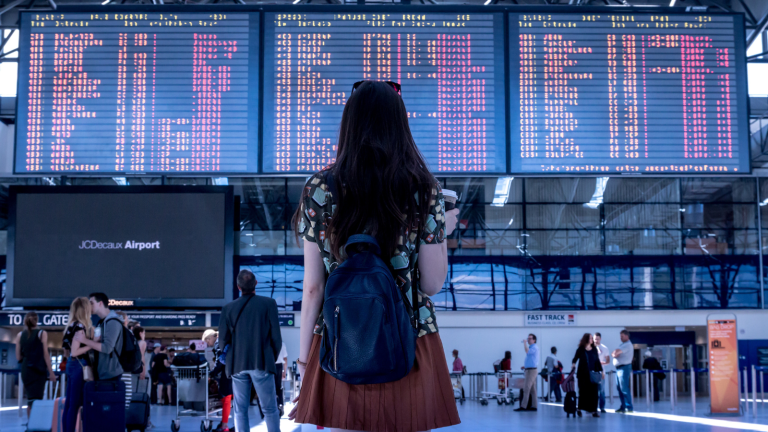 A woman with long hair is standing inside an airport terminal, looking up at a large flight information board displaying departures. She has a blue backpack, and people with luggage are visible around her.