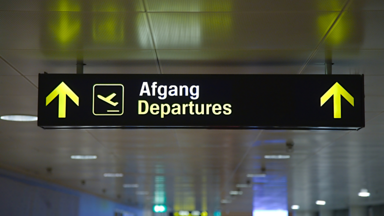 A sign in an airport terminal with yellow arrows pointing up. It displays the words Afgang and Departures alongside an airplane icon, guiding travelers to the departure area. The background is a ceiling with lights.