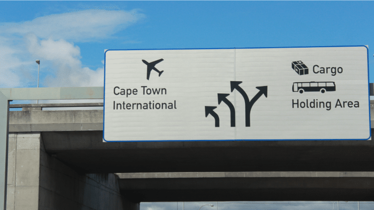 A road sign on a highway indicating directions. The sign displays arrows for Cape Town International with an airplane symbol, and Cargo Holding Area with a bus and package icon. It is mounted on a concrete overpass under a partly cloudy sky.