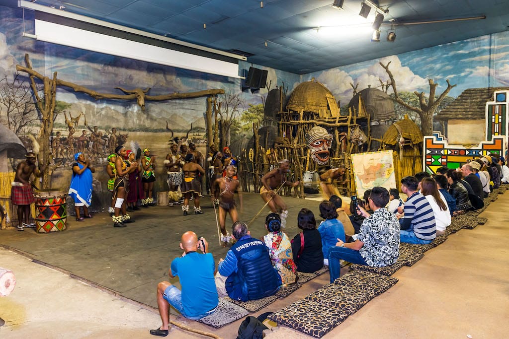 Dancers in traditional attire perform energetically on a stage with a village backdrop, reminiscent of Lesedi Cultural Village, as an audience sits on benches watching the cultural show. The scene includes colorful costumes and drums, creating a vibrant and lively atmosphere.