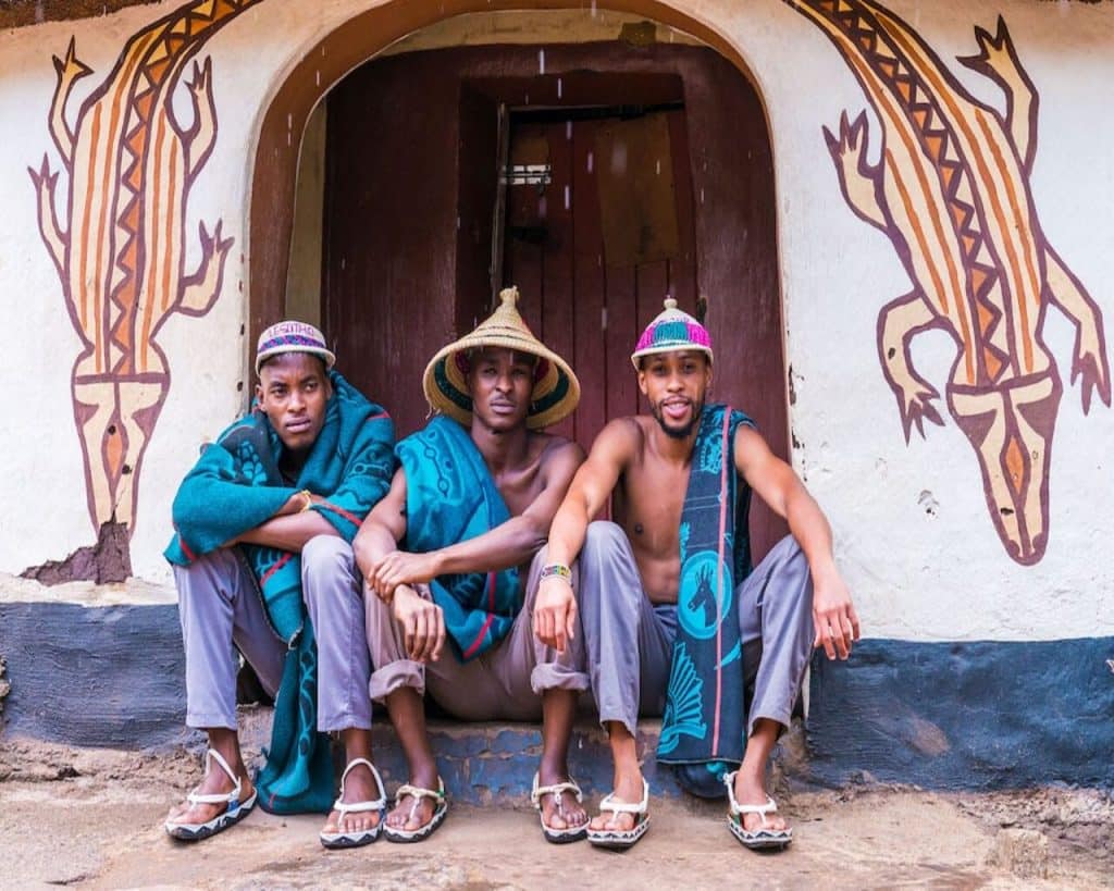 Three men in traditional hats and colorful fabrics sit on a step before a decorated building at Lesedi Cultural Village. The intricate animal designs on the wall frame their relaxed presence, vividly showcasing cultural attire and heritage amidst this vibrant community hub.