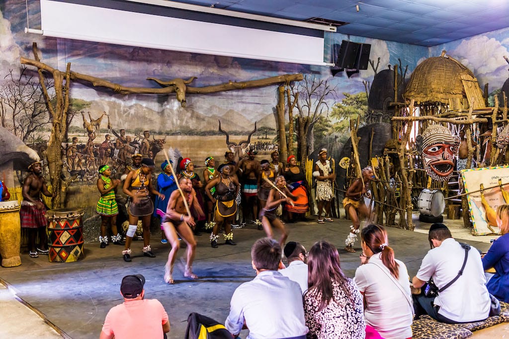 A group of people performs a traditional dance in brightly colored attire, reminiscent of the vibrant displays at Lesedi Cultural Village. The backdrop features an African village scene with huts and a mural. Seated onlookers enjoy the captivating performance in an indoor setting.