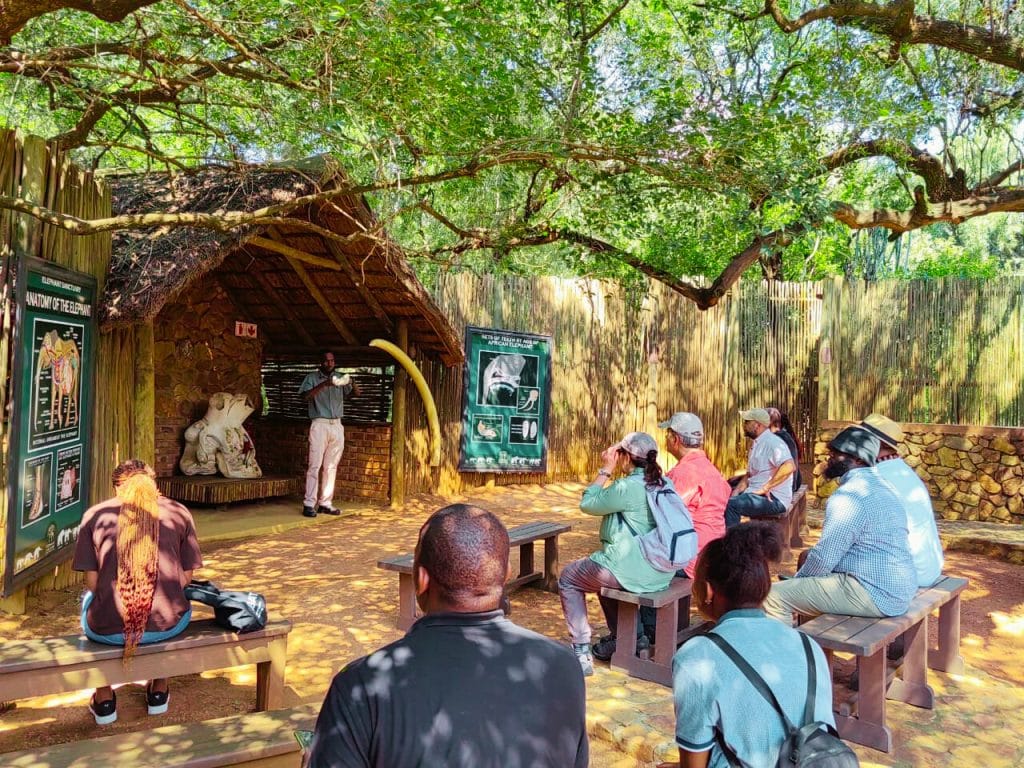 A group of people sits on benches under a tree, listening to a guide at the elephant sanctuary. The scene is outdoors, with informational posters displayed on a rustic wooden shelter. The atmosphere is casual and educational.