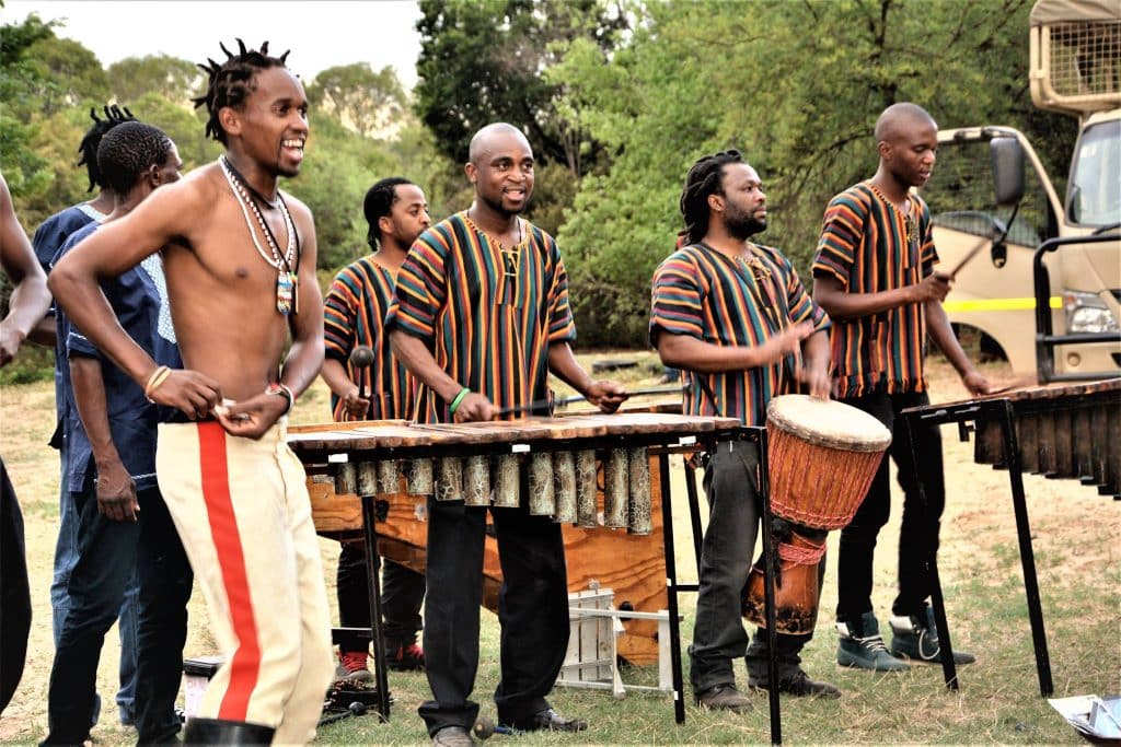 A group of musicians delights visitors at the lion park tour, performing outdoors with several men playing marimbas and one on a drum. They wear colorful striped shirts, adding vibrant charm to the scene, surrounded by lush greenery and a parked vehicle in the background.