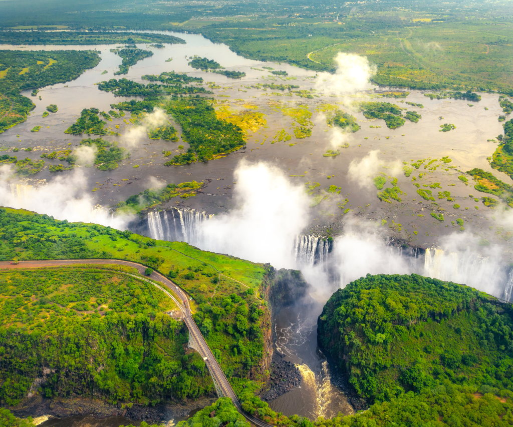 Aerial view of a large waterfall surrounded by lush greenery and mist. The river flows over a wide cliff, creating a striking cascade. A road with a bridge curves through the foreground, while clouds drift above.