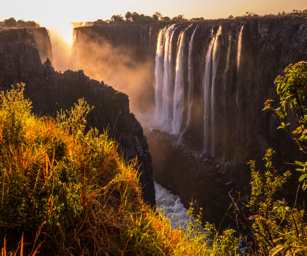 A stunning view of a waterfall cascading over a rocky cliff at sunset, surrounded by lush green foliage. The golden light creates a mist, adding a warm glow to the scene.