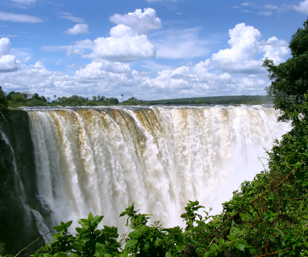 A powerful waterfall cascades over a wide cliff under a bright blue sky with scattered clouds. Lush green foliage surrounds the foreground, framing the view of the majestic waterfall.