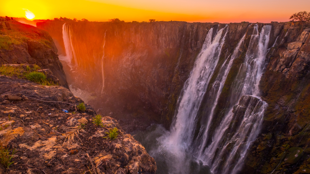 A breathtaking view of a large waterfall cascading into a gorge at sunset. The sky glows orange, and the surrounding cliffs are bathed in warm light, highlighting the lush greenery on the rocky edge.