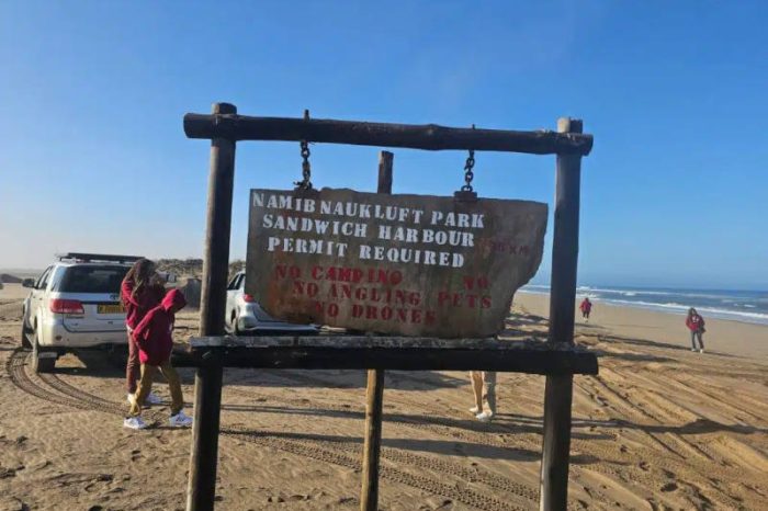 A weathered wooden sign at the sandy entrance reads Namib Naukluft Park, Sandwich Harbour, Permit Required. It also warns: No camping, No angling, No pets, and No drones. In the background, people and a vehicle highlight this scenic spot on a Namibia tour from Johannesburg.