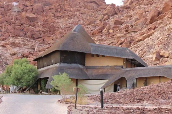 A thatched-roof building nestled in a rugged, rocky landscape with reddish-brown hues evokes the charm of a Namibia tour from Johannesburg. The structure has multiple levels and sloped roofs, harmonizing with nature. A small tree and a paved path grace the foreground.