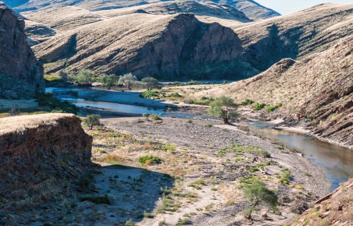 A winding river flows through a rocky canyon with dry, grassy hills on both sides under a clear blue sky. Sparse vegetation lines the riverbanks, creating a striking contrast with the rugged landscape—a scene reminiscent of a Namibia tour from Johannesburg.