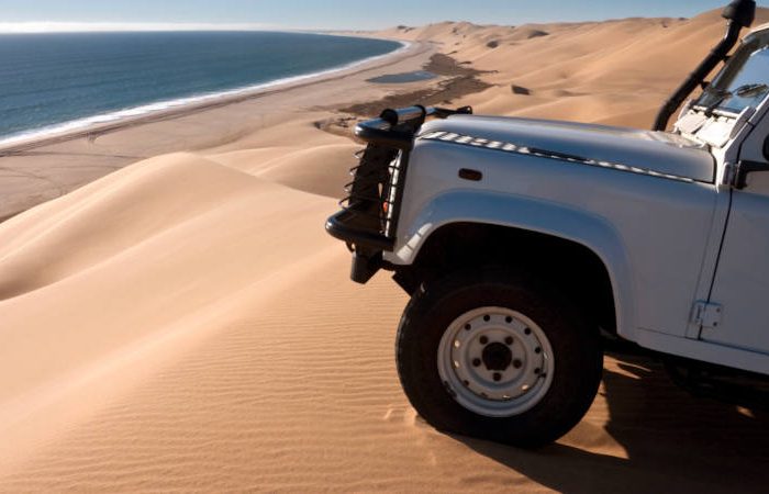 A white off-road vehicle, part of a Namibia tour from Johannesburg, is parked on a sand dune overlooking a scenic coastline. The view includes vast sand dunes merging into the blue ocean under a clear sky.