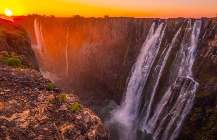 Experience the spectacular sight of a waterfall reminiscent of Namibia's rugged beauty, plunging down a steep cliff at sunset. The sky, painted with warm orange and pink hues, illuminates the mist and water cascading over the rocks, akin to the breathtaking Victoria Falls.