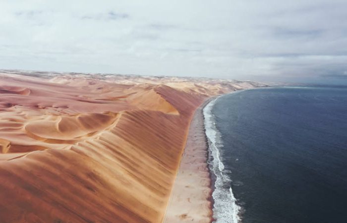 Aerial view of a dramatic landscape where desert sand dunes meet the ocean. The orange dunes stretch along the coast, contrasting with the deep blue sea under a cloudy sky—a stunning scene typical of a Namibia tour from Johannesburg.