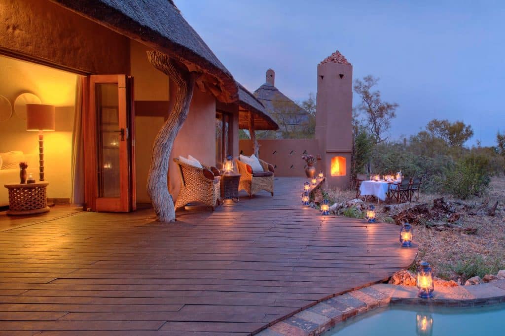 Rustic lodge patio at sunset with wicker chairs and a small pool. Candlelit lanterns line the wooden deck leading to a dining table set for two. Bushland and a tower with a fireplace are visible in the background.