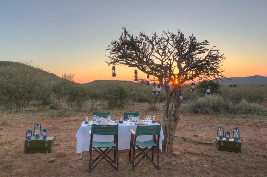 A serene outdoor dining setup at sunset in a savannah landscape. Four chairs surround a table with a white tablecloth and tableware. A tree decorated with lanterns stands nearby, with the sun setting in the background.