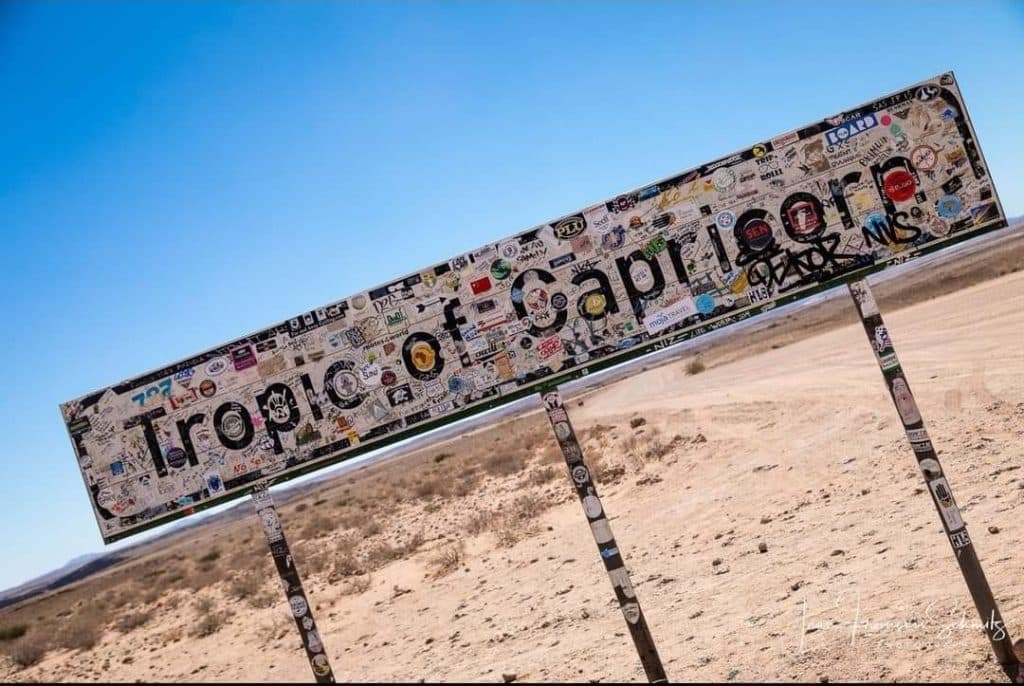 A metallic sign reading Tropic of Capricorn, covered in stickers and graffiti, marks a stop in the dry, barren landscape of Namibia. Under a clear blue sky, it's a popular photo spot for those on the Namibia tour from Johannesburg.