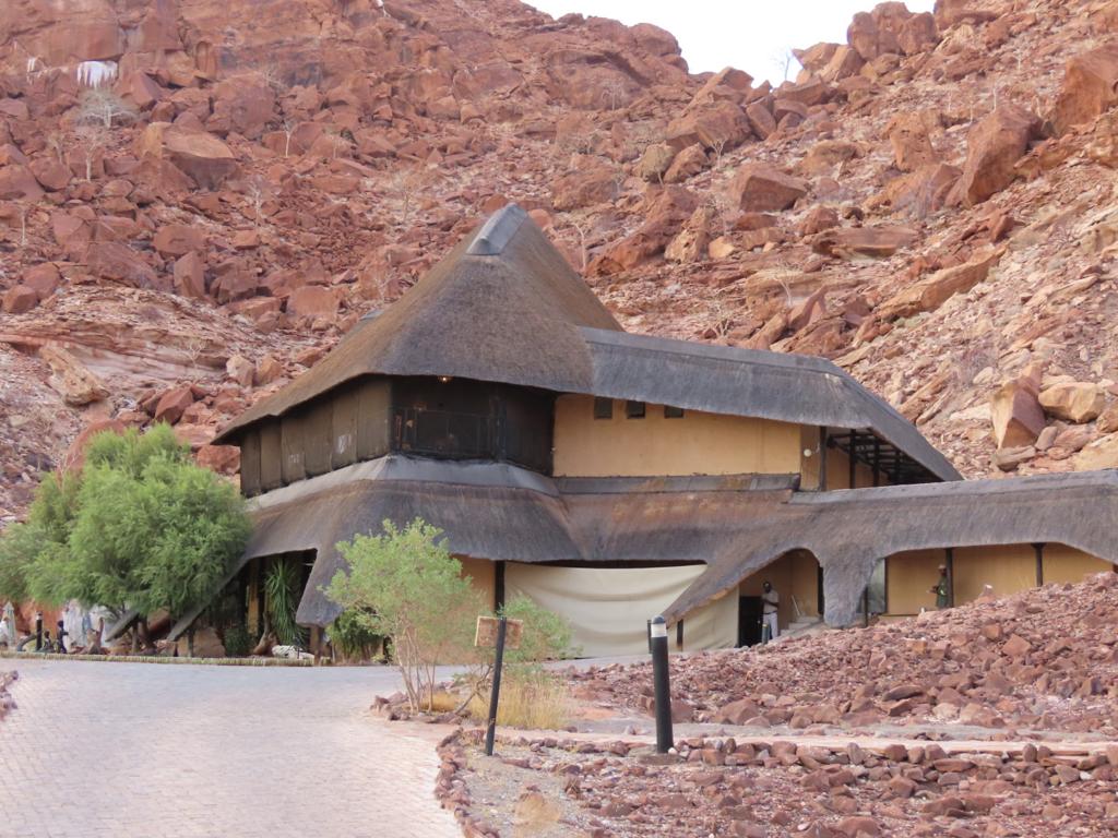 A thatched-roof building nestled in a rugged, rocky landscape with reddish-brown hues evokes the charm of a Namibia tour from Johannesburg. The structure has multiple levels and sloped roofs, harmonizing with nature. A small tree and a paved path grace the foreground.