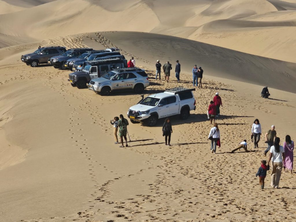 A group of people gather around several SUVs parked on a sandy dune in the desert landscape, part of an exhilarating Namibia tour from Johannesburg. Footprints trace their path as some walk or kneel, while others stand near the vehicles, soaking in the adventure.