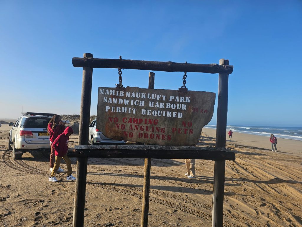 A weathered wooden sign at the sandy entrance reads Namib Naukluft Park, Sandwich Harbour, Permit Required. It also warns: No camping, No angling, No pets, and No drones. In the background, people and a vehicle highlight this scenic spot on a Namibia tour from Johannesburg.