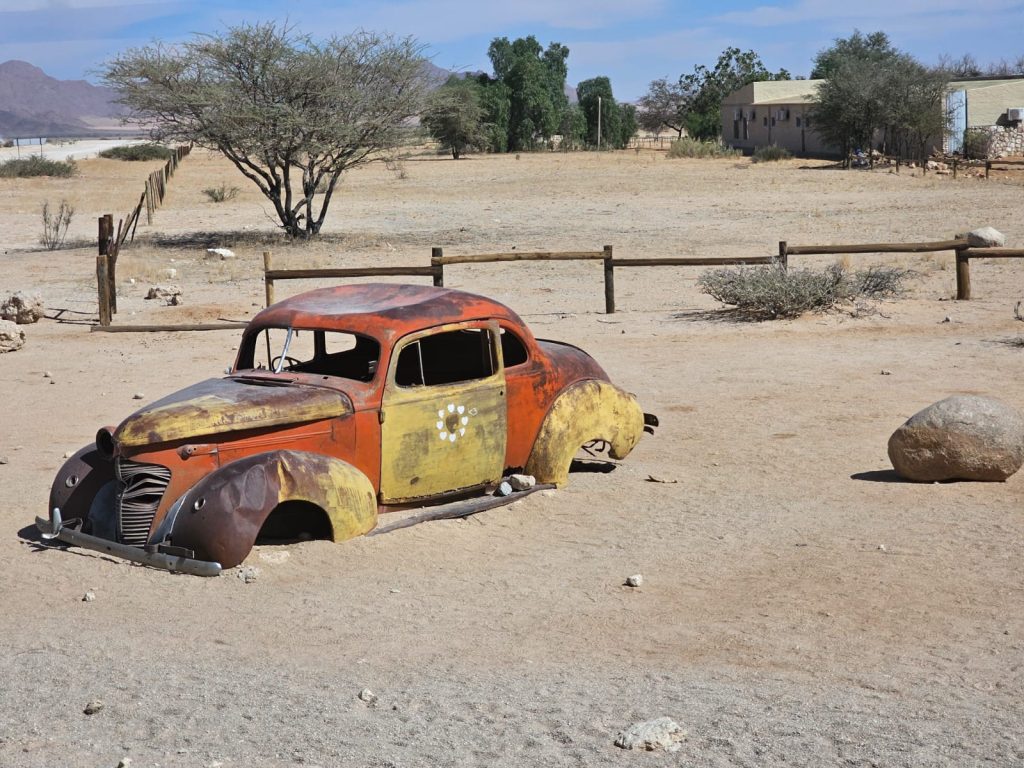 A rusted, vintage car is partially buried in a sandy landscape typical of a Namibia tour from Johannesburg. The dry area has sparse vegetation, with a few trees and shrubs dotting the scenery. A small fence and building are visible in the background under the clear blue sky.