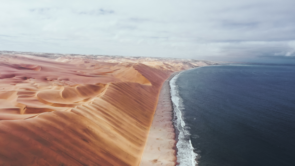 Aerial view of a dramatic landscape where desert sand dunes meet the ocean. The orange dunes stretch along the coast, contrasting with the deep blue sea under a cloudy sky—a stunning scene typical of a Namibia tour from Johannesburg.