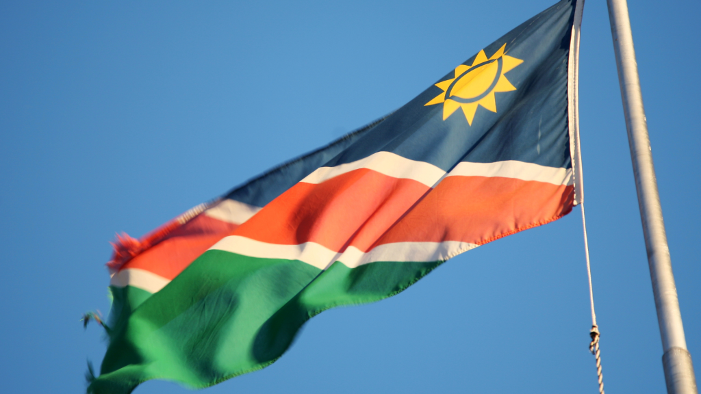 Against the backdrop of a clear blue sky, the flag with its blue, red, and green horizontal stripes and white border waves proudly. The yellow sun with 12 triangular rays on the blue stripe shines brightly, reminiscent of embarking on a Namibia tour from Johannesburg.