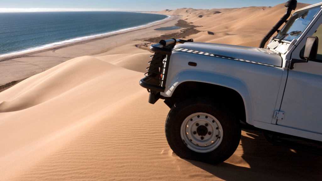 A white off-road vehicle, part of a Namibia tour from Johannesburg, is parked on a sand dune overlooking a scenic coastline. The view includes vast sand dunes merging into the blue ocean under a clear sky.