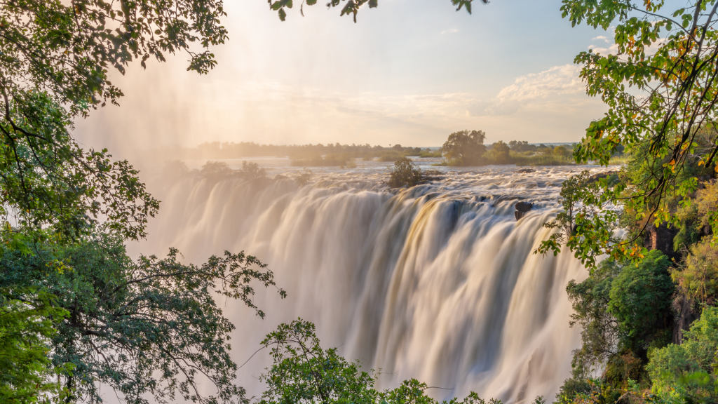 A scenic view of Victoria Falls cascading over a cliff, surrounded by lush green foliage reminiscent of Namibia's landscapes. The partly cloudy sky and sunlight create a misty atmosphere around the waterfall, adding a golden hue to the scene.