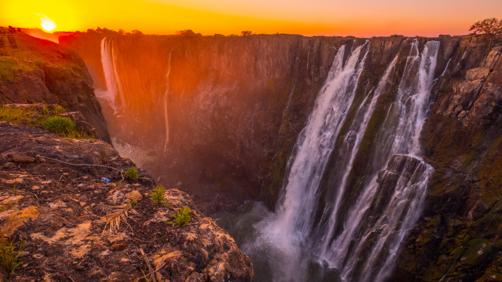 Experience the spectacular sight of a waterfall reminiscent of Namibia's rugged beauty, plunging down a steep cliff at sunset. The sky, painted with warm orange and pink hues, illuminates the mist and water cascading over the rocks, akin to the breathtaking Victoria Falls.