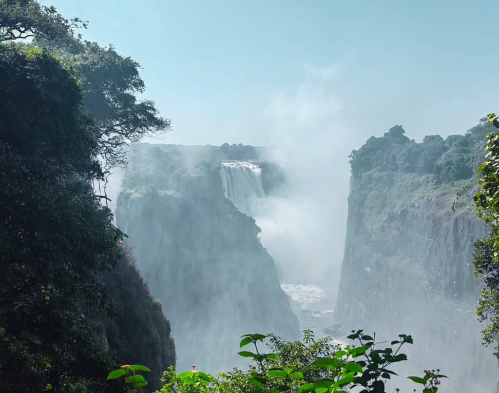 A majestic waterfall cascades between rocky cliffs, surrounded by lush green foliage. Mist rises from the base, creating a dreamy atmosphere under a clear blue sky.