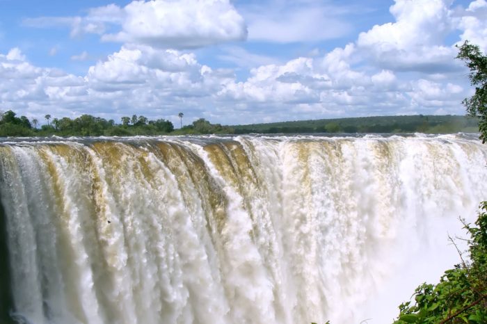 A powerful waterfall cascades over a wide cliff under a bright blue sky with scattered clouds. Lush green foliage surrounds the foreground, framing the view of the majestic waterfall.