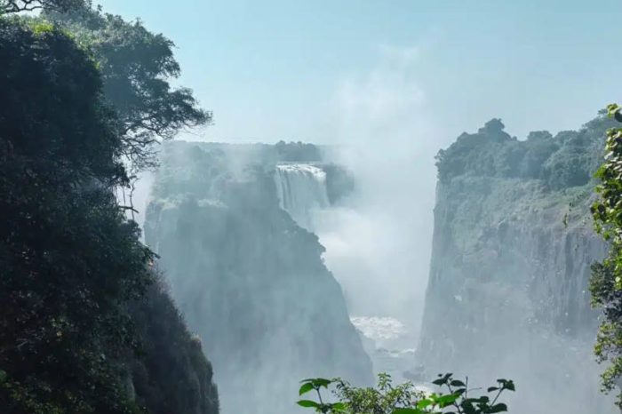 A majestic waterfall cascades between rocky cliffs, surrounded by lush green foliage. Mist rises from the base, creating a dreamy atmosphere under a clear blue sky.