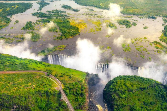 Aerial view of a large waterfall surrounded by lush greenery and mist. The river flows over a wide cliff, creating a striking cascade. A road with a bridge curves through the foreground, while clouds drift above.