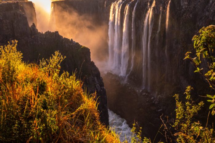 A stunning view of a waterfall cascading over a rocky cliff at sunset, surrounded by lush green foliage. The golden light creates a mist, adding a warm glow to the scene.