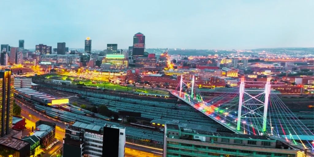 A vibrant cityscape at dusk featuring a well-lit, colorful cable-stayed bridge crossing railway tracks. The skyline is dotted with illuminated buildings under a clear, fading sky.