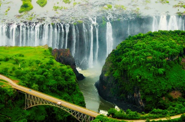 Aerial view of a waterfall cascading down a cliff with lush green surroundings. A bridge crosses the river at the base, surrounded by vibrant greenery. The scene is vibrant and dynamic, showcasing the waterfalls power and beauty.