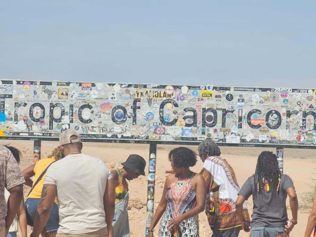 A group of people stand in front of a large sign covered in stickers that reads Tropic of Capricorn in a desert landscape under a clear blue sky. The people are casually dressed, and the atmosphere is bright and sunny.