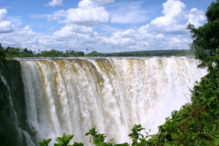 A powerful waterfall cascades over a wide cliff under a bright blue sky with scattered clouds. Lush green foliage surrounds the foreground, framing the view of the majestic waterfall.