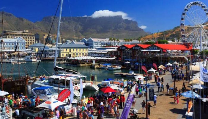 A lively waterfront bustling with tourists exploring Cape Town tour packages, boats docked in a harbor, and a large Ferris wheel. The scene is set against majestic mountains under a clear blue sky, with vibrant shops and people enjoying the day.