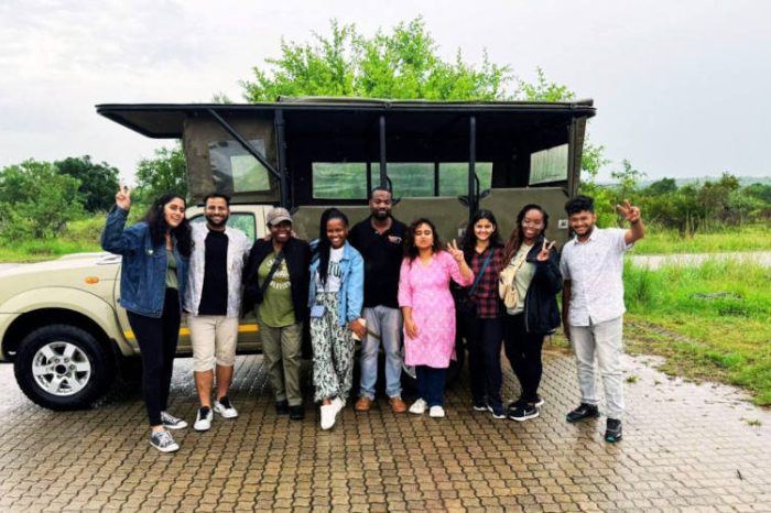 A group of people smiling and posing in front of an open safari vehicle, embarking on their 2-day Kruger safari from Johannesburg. One person is holding up a peace sign amid the lush greenery. The sky is overcast, adding to the anticipation of adventure.