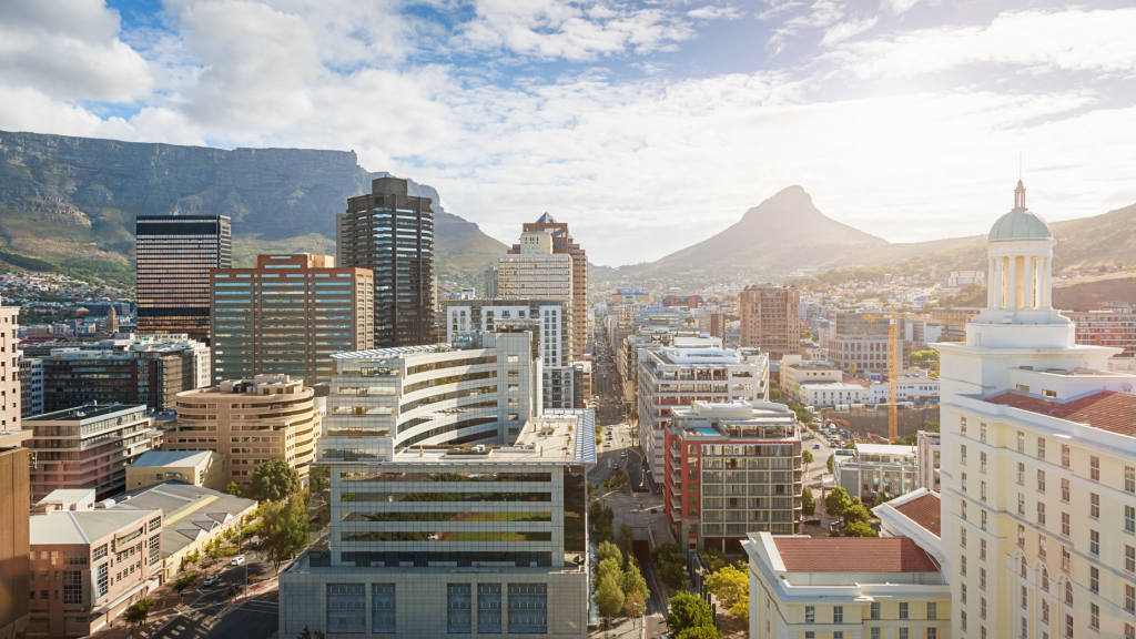 Aerial view of a modern cityscape with tall buildings, including a domed structure. In the background, a majestic mountain range is illuminated by the sun, with a partly cloudy sky overhead.